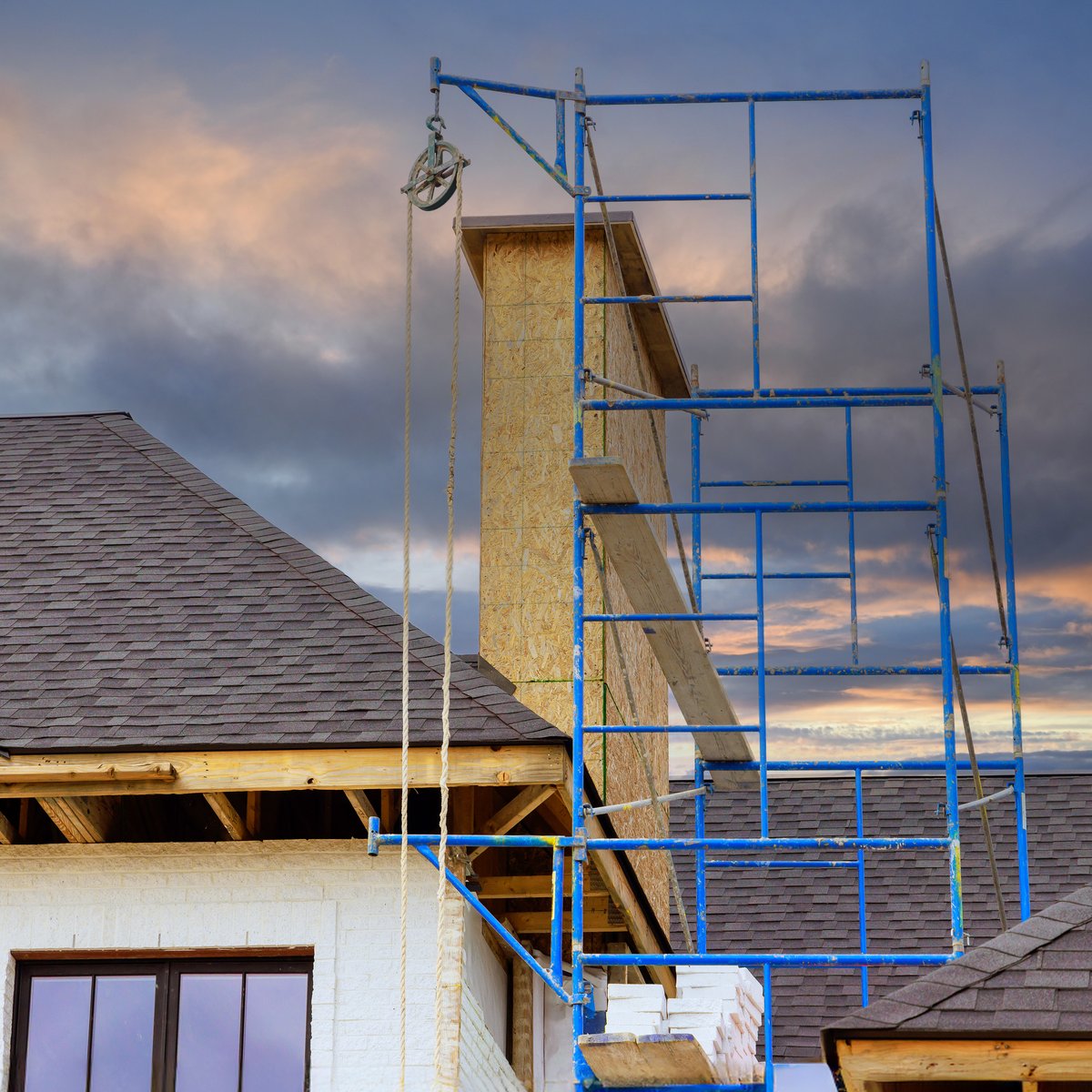 Chimney stripped to sheathing and wrapped in blue scaffolding above a shingle roof at dusk - chimney rebuild in Chicago, IL by MasonryPros.us