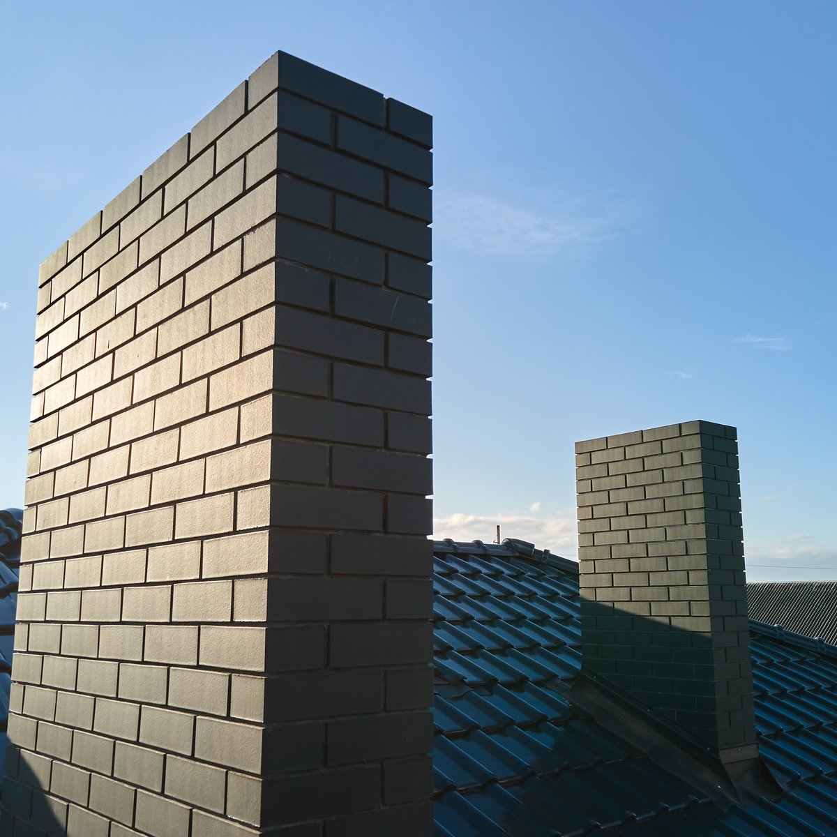 Two grey brick chimney stacks rising above a tile roof against a clear sky - chimney tuckpointing and repointing in Chicago, IL by MasonryPros.us